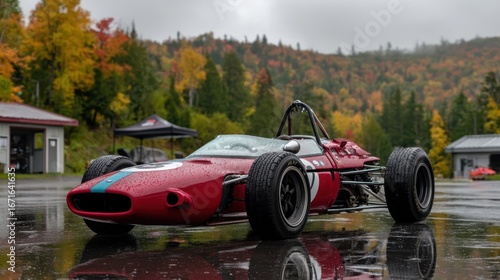 A classic red race car sits on a wet track surrounded by autumn trees and misty hills in the background