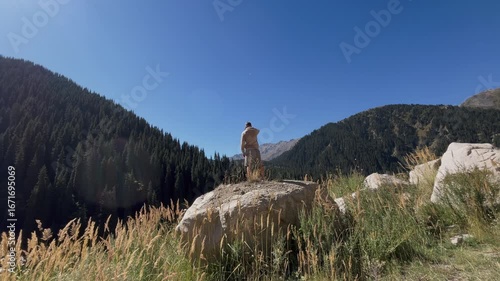 Man climbs onto a huge stone against the backdrop of scenic mountains and coniferous forest. Outdoor adventure scene with wilderness landscape, rocky terrain, and natural beauty of untouched highland 