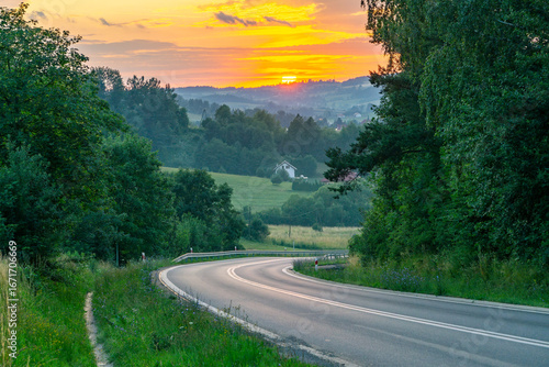 Fototapeta Naklejka Na Ścianę i Meble -  Curved asphalt road in the Bieszczady Mountains at sunset, picturesque valley with houses and green hills.
