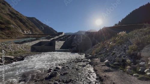 Huge concrete dam stands on a mountain river with flowing water. Morning sunlight shines over the peaks in the background, combining industrial architecture, renewable energy concept, and scenic natur