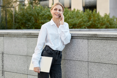 A young blonde woman in a white blouse and black jeans goes to an interview with a laptop in her hands