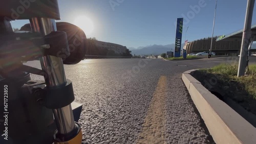 Camera mounted on moving transport captures drive along city road. Urban traffic scene with cars, asphalt, and street infrastructure. In the distance, mountain range rises above the city, combining ur