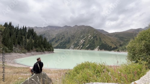 Panoramic view of a mountain lake surrounded by rocky slopes. A lone person sits on a stone, observing the scenic landscape. Peaceful nature scene with calm water, mountains, and dramatic cloudy sky.
