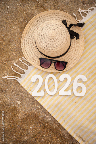 Stylish beach accessories like a straw hat, sunglasses, and a striped towel rest on sandy shores, with wooden numbers 2026 hinting at upcoming beach vacations