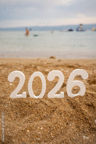 White numbers reading 2026 resting on sandy beach, with a blurred sea and people enjoying the water in the background, capturing the essence of upcoming New Year celebrations