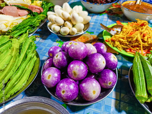Close-up of small purple eggplants or terong bulat unggu in a full bowl with a variety of fresh and boiled vegetables such as winged beans, okra, and white eggplants, for eating with chili paste.