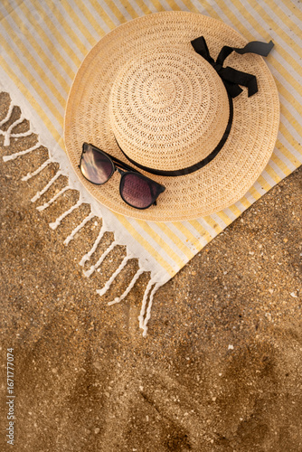 Beach towel, straw hat, and sunglasses lying on sand suggest a relaxing summer day at the beach, inviting viewers to envision a carefree vacation