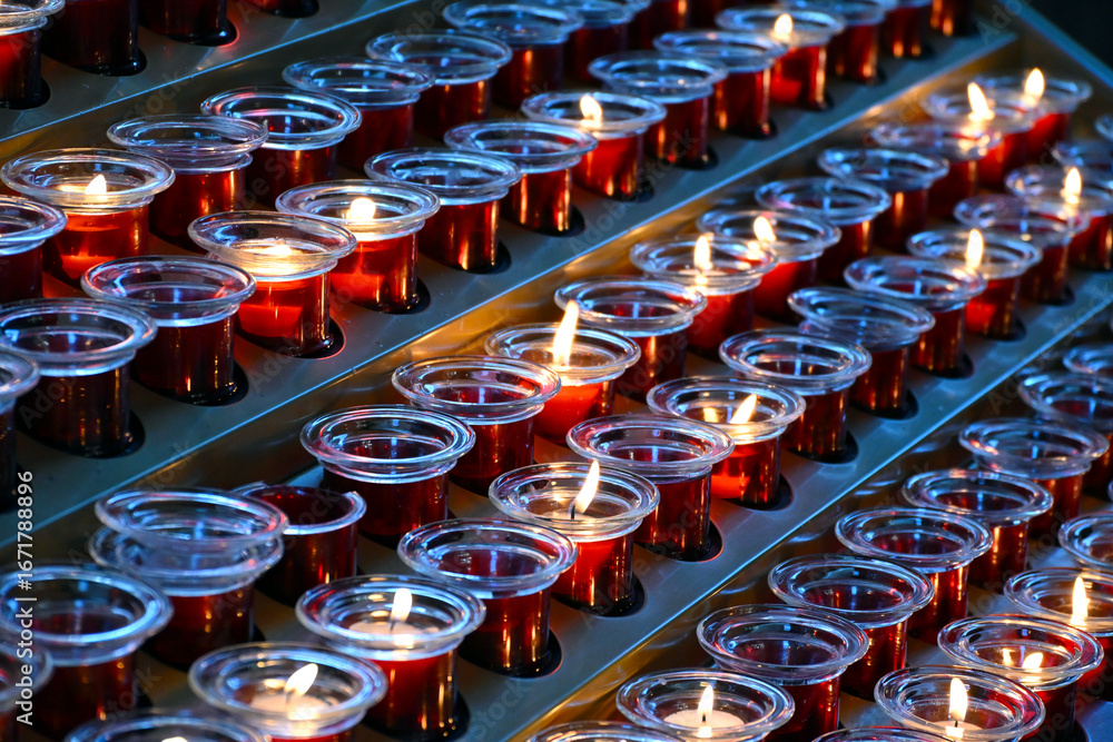 Fototapeta premium Small votive candles, lighted and placed in red glass candlesticks among empty ones, interior details of a catholic church