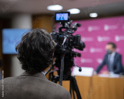 Person with curly hair is seen from behind, operating video camera during press conference. setting features professional backdrop with signage, indicating formal event