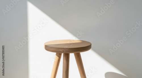 Simple round wooden stool standing in a beam of bright natural sunlight creating a strong shadow on a white wall.