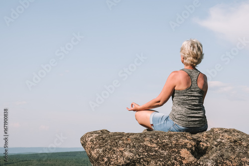 woman sits cross-legged on large rock, meditating peacefully while enjoying panoramic view vast green hills under bright blue sky. rear view. close up.