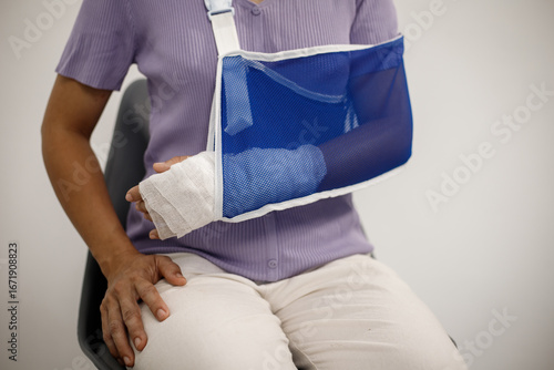 A person from the chest up wears a light green cardigan over a white shirt, with their right forearm wrapped in a beige bandage and supported inside a blue mesh arm sling.