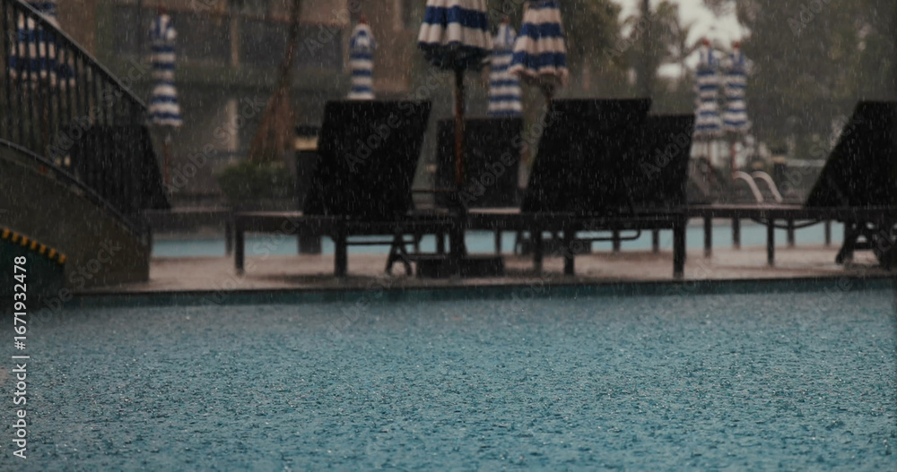 Fototapeta premium Rainy day at outdoor swimming pool with loungers and umbrellas, creating tranquil atmosphere. Captures relaxation, leisure, and nature's beauty in urban resort setting.