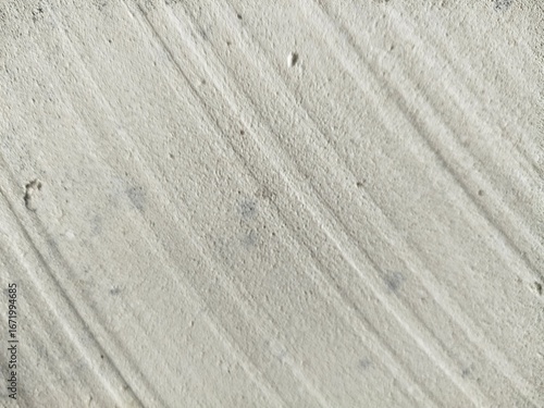 * A close-up shot of a rough, light gray concrete or cement surface. The texture is prominently featured, with parallel diagonal lines or grooves etched into the material. 