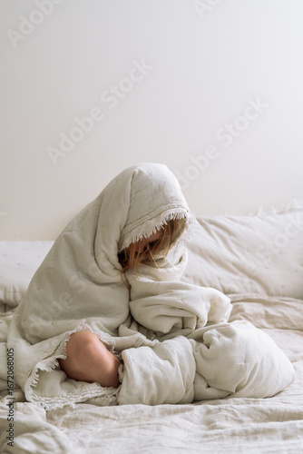 Woman or teenage girl wrapped in a linen blanket, sitting on a bed in a calm, minimalist bedroom. Soft light and cozy textures create a peaceful morning mood. beauty of slow mornings. Bed rotting