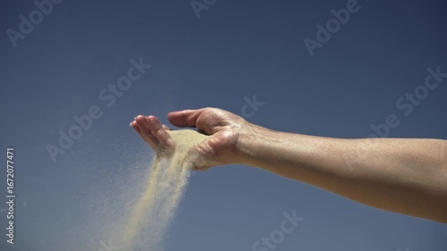 Slow-motion close-up of a woman's hand with sand slipping through fingers against a sky background. Symbolizes fleeting time and life's transience.