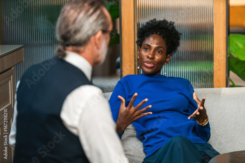 Two professionals on a sofa in a modern office, engaged in a focused conversation. One gestures while explaining, suggesting coaching, mentoring, or consulting.