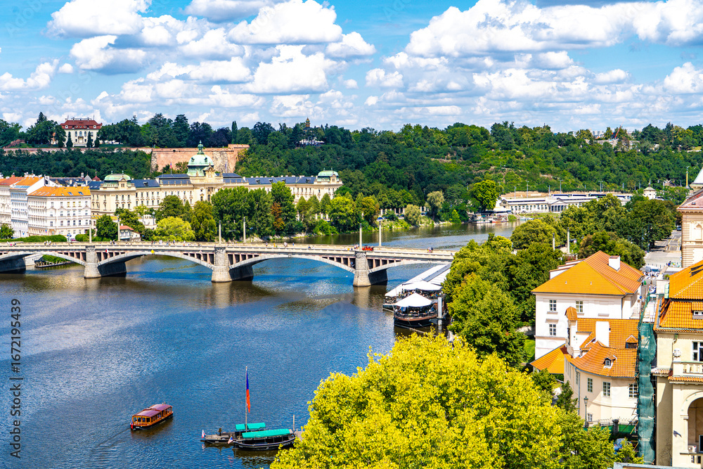 Obraz premium he Vltava River, surrounded by lush summer greenery, with a bridge connecting the historic districts of the Old Town under a blue sky with clouds.Prague,Czech Republic