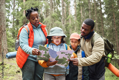 Black woman and Black man guiding two children while reading map together in forest, family group exploring nature, all focused on navigation during outdoor adventure