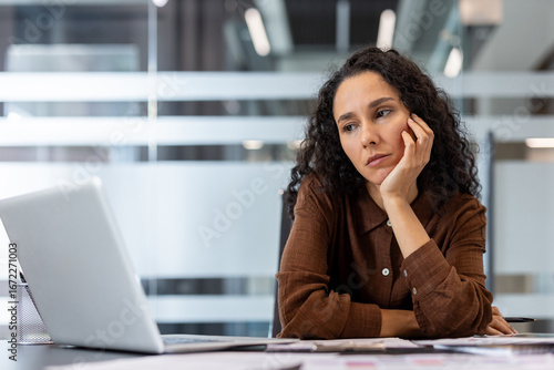 Tired professional woman looking at a laptop in a modern office.