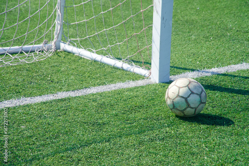 Картината върху платно Old soccer ball by the goalpost on green artificial turf, right on the goal line — a tense near‑miss at the corner of the net in bright sun