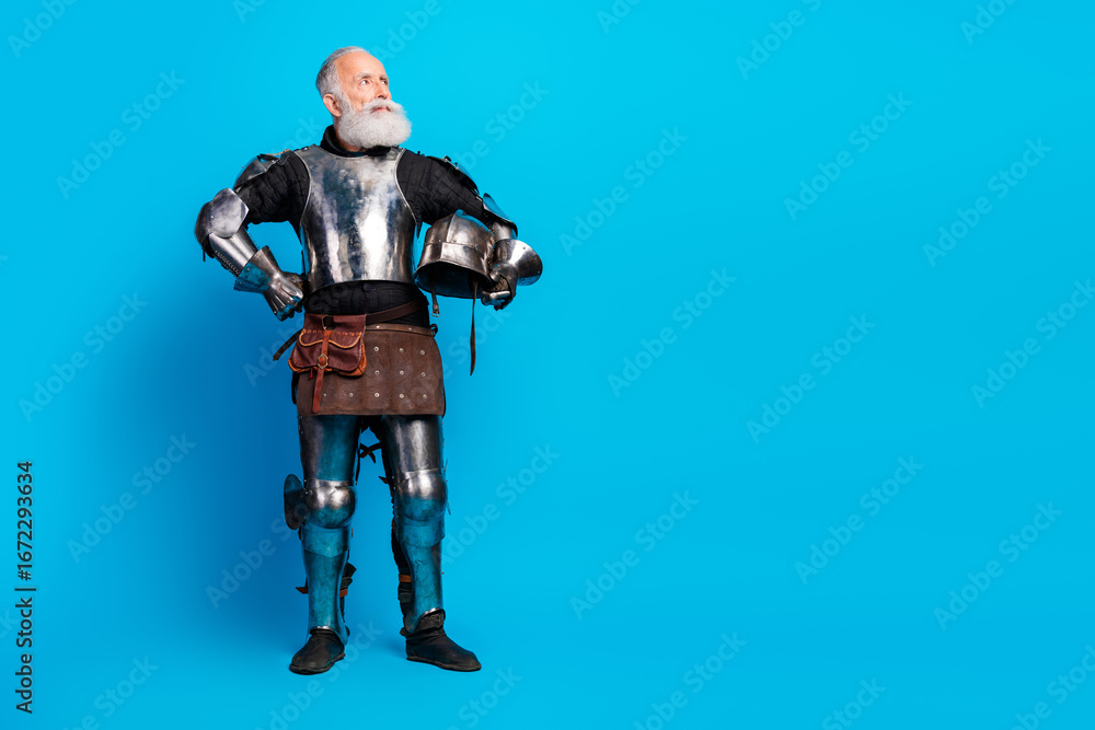 Naklejka premium Senior man dressed in medieval knight armor holding a helmet against a blue background