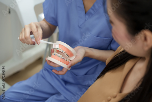Asian female dental assistant demonstrating correct brushing technique on orthodontic model to adult patient. Explanation focused on oral hygiene and dental maintenance after braces treatment.