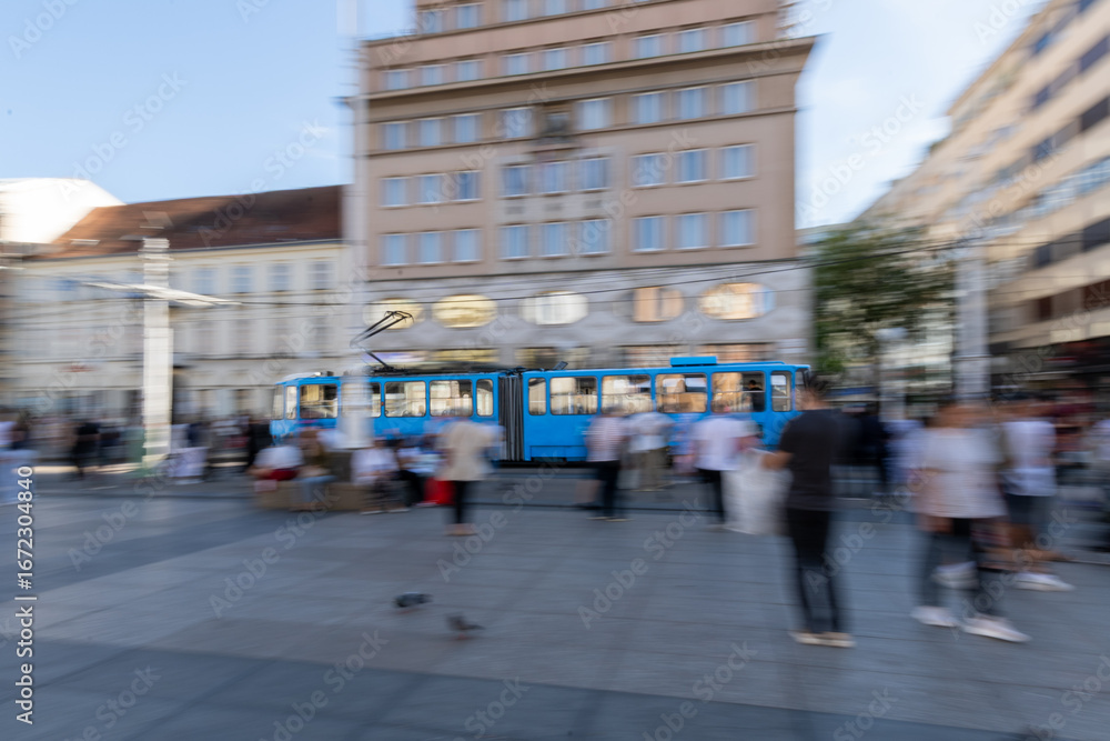 Obraz premium A blue tram moves through Zagreb's lively city square, surrounded by modern and historic buildings and a bustling crowd. motion and panning effect