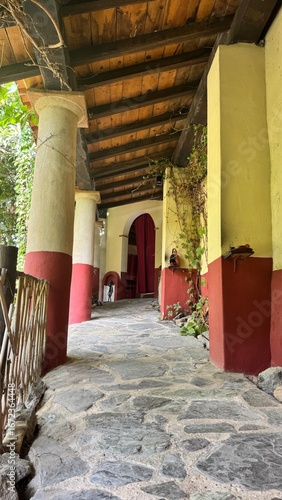 Traditional Roman Villa Architecture: Classic Colonnade Path with Red and Yellow Columns, Stone Pavement and Wooden Roof in an Ancient Mediterranean Garden Setting