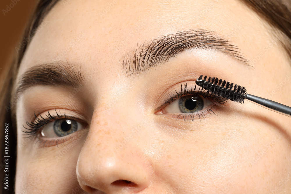Naklejka premium Woman applying mascara onto her eyelashes on brown background, closeup