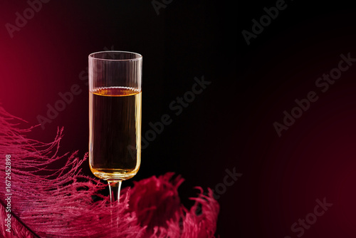 Stylish flute glass with sparkling wine or champagne, surrounded by red feather details on dark luxury background. Beautiful glamorous romantic greeting card