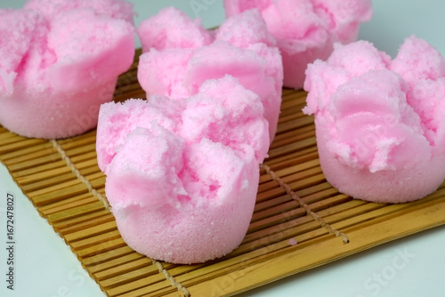 A close-up of a vibrant pink Kue Mangkok or Huat Kueh, a traditional steamed sponge cake from Southeast Asia with a signature cracked top. The cakes are on a bamboo mat with a plain background.