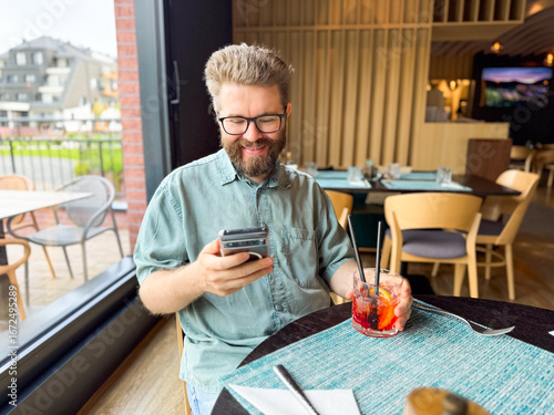 Man sitting at restaurant table holding smartphone and cocktail. Leisure, lifestyle, and hospitality atmosphere with social interaction and guest experience.
