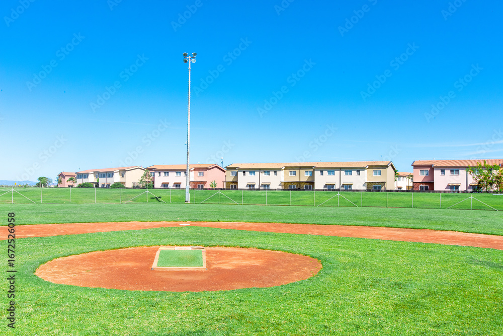 Obraz premium A baseball field with a wide garden surrounding it, offering a view of nearby houses in the distance. The lush green field contrasts with the neighborhood, creating a peaceful, suburban atmosphere.