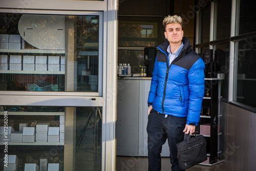 Wallpaper Mural Handsome young man exits modern building wearing blue puffer jacket with backpack and briefcase on a sunny day Torontodigital.ca