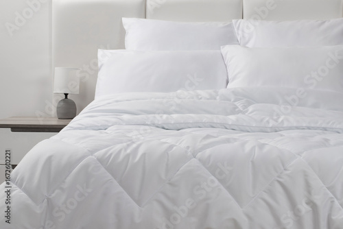 Foto A bed with white pillows and a blanket, next to a bedside table with a lamp, day