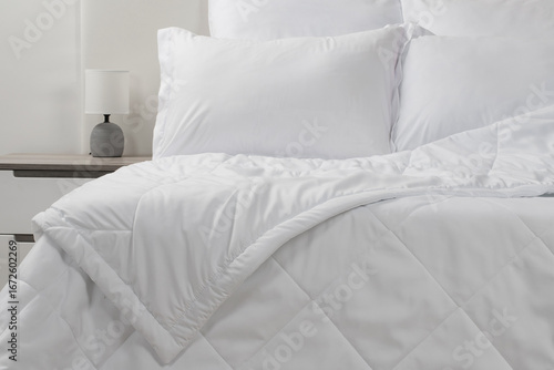 A bed with white pillows and a blanket, next to a bedside table with a lamp, daylight