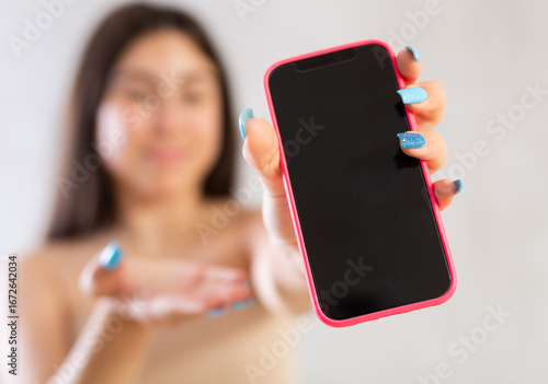Smartphone with dark screen held by young woman demonstrating it standing against light unicolored wall