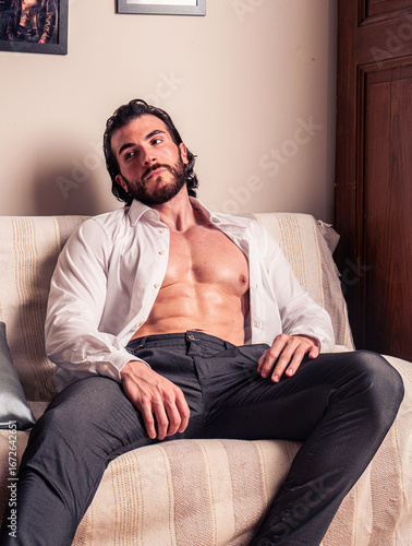 A young man with dark hair and a beard sits on a comfortable couch, dressed in an unbuttoned white shirt, looking thoughtfully toward the side in a cozy living room