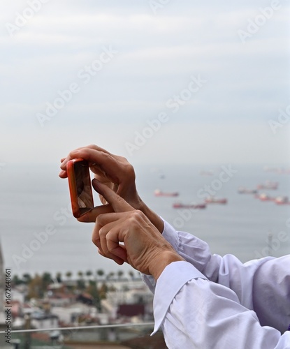 Individual holding smartphone with a scenic view of the ocean and ships in the background, capturing a moment of tranquility and connection with nature