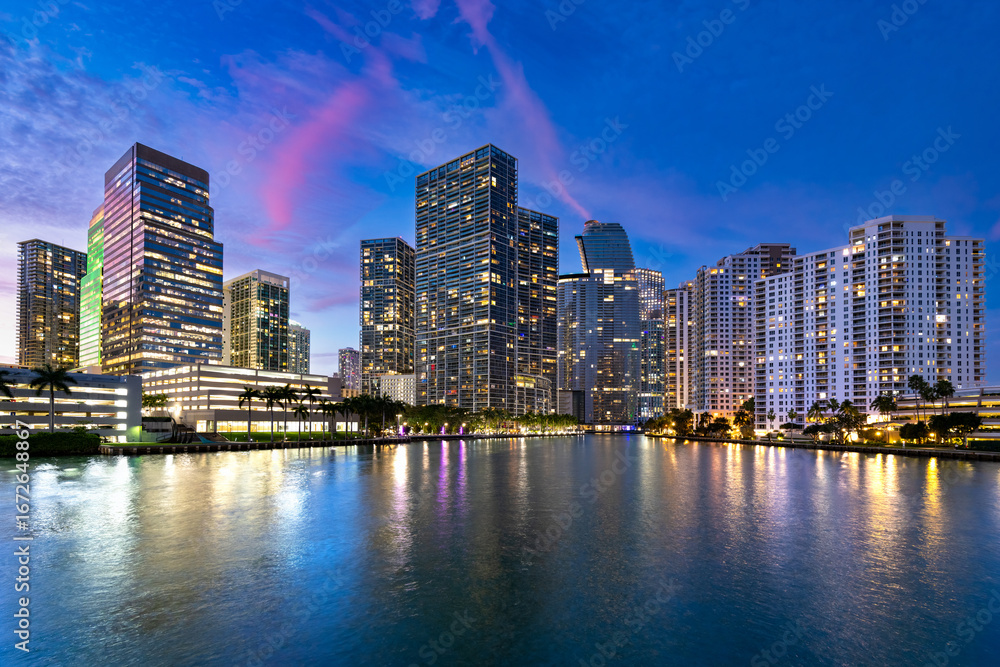 Obraz premium The vibrant skyline of the Brickell neighborhood in Miami, with illuminated buildings and palm trees under a colorful twilight sky.