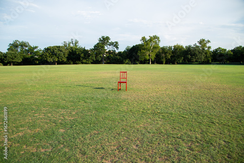 A single red chair stands in the middle of a wide grassy field under a clear sky. Symbolic and artistic, representing solitude, minimalism, and creativity.