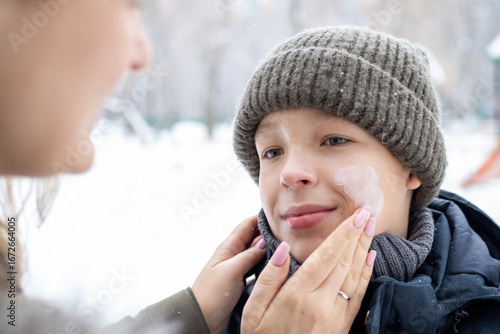 Caring mother applying protective sunscreen or moisturizing cream on her son's face during wintertime outdoor activities