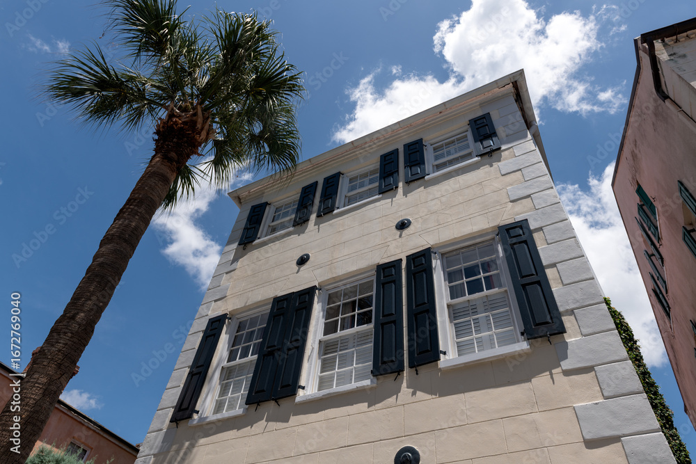 Fototapeta premium Charleston, South Carolina, USA - July 2, 2025: Low-angle view of a historic Charleston stucco house with black shutters and palm tree under a bright blue sky.