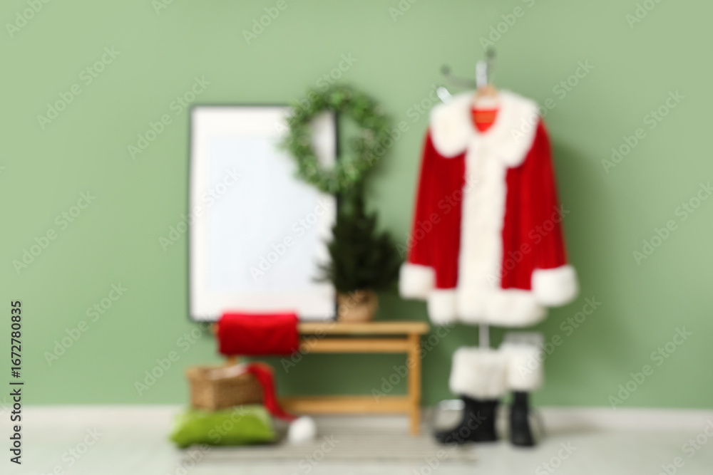 Fototapeta premium Hanger with Santa Claus suit, bench and frame near green wall. Blurred view
