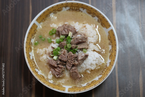A close-up of a (coto makassar) delicious beef rice bowl with tender slices of beef, topped with fresh green onions, served in a flavorful broth.