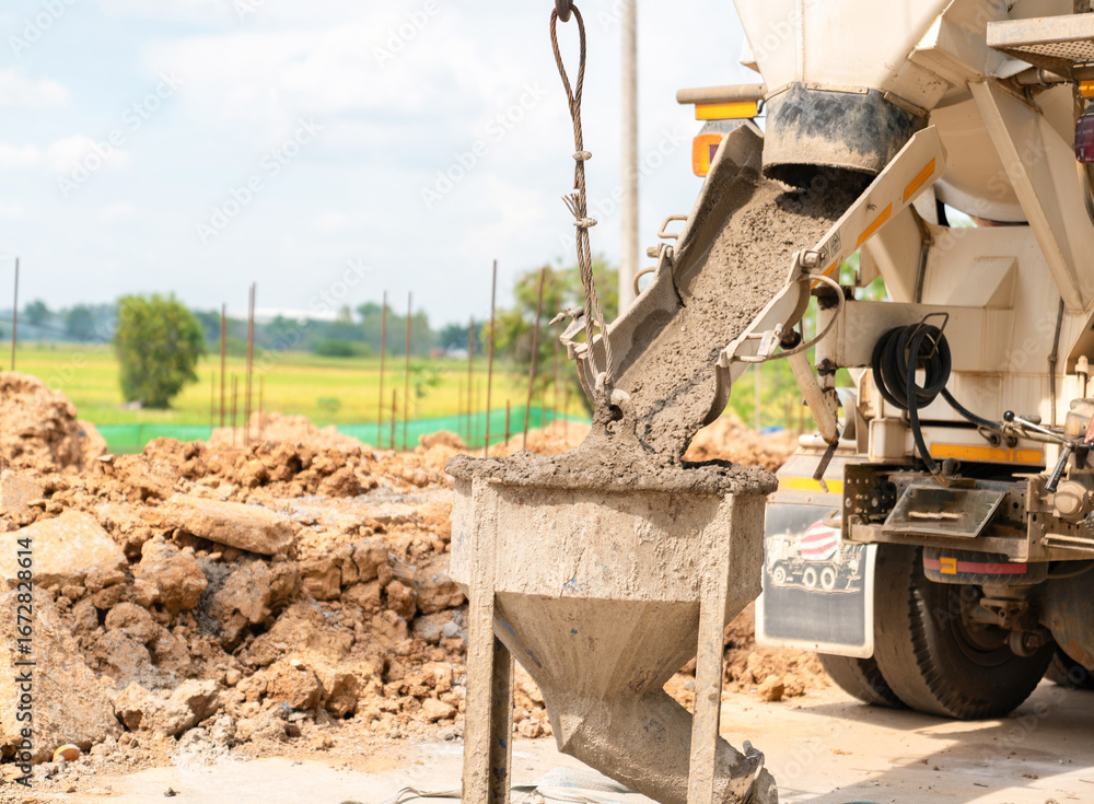 Fototapeta premium Concrete being poured from a cement mixer truck into a metal bucket at a construction site in a rural area.