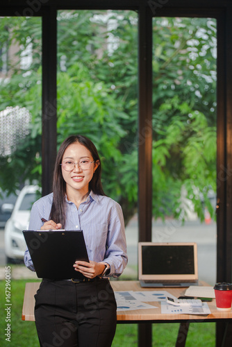Smiling young Asian businesswoman is thinking and analyzing work plan in office hard using laptop, smartphone, calculator and documents. Chart sheet on table. Business and finance concept.