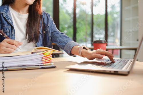 Smiling young Asian woman clearing paperwork in office, using laptop and many documents beside her. Accounting and finance business concept.