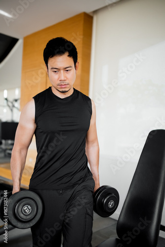 Asian man standing beside bench holding dumbbells in gym